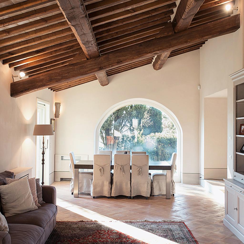 Rustic living and dining area with exposed wooden beams, arched window, and cozy furnishings, designed by Archidomus and Architetto Monica Rossi.