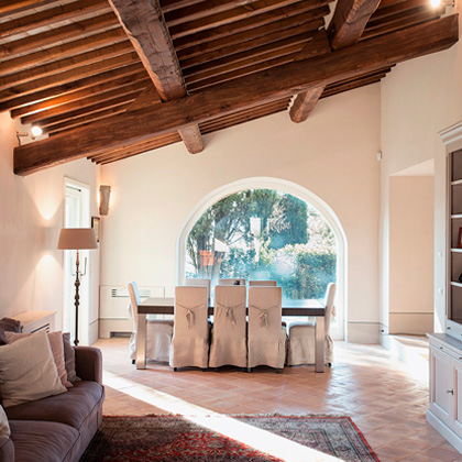 Rustic living and dining area with exposed wooden beams, arched window, and cozy furnishings, designed by Archidomus and Architetto Monica Rossi.