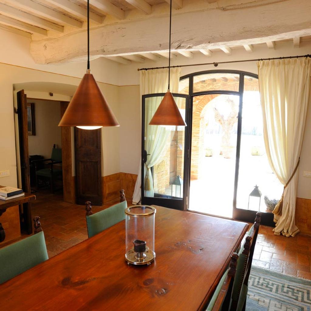 Sala da pranzo con tavolo in legno, lampade a sospensione in rame e vista sull'esterno, progettata da Archidomus architetto Monica Rossi.